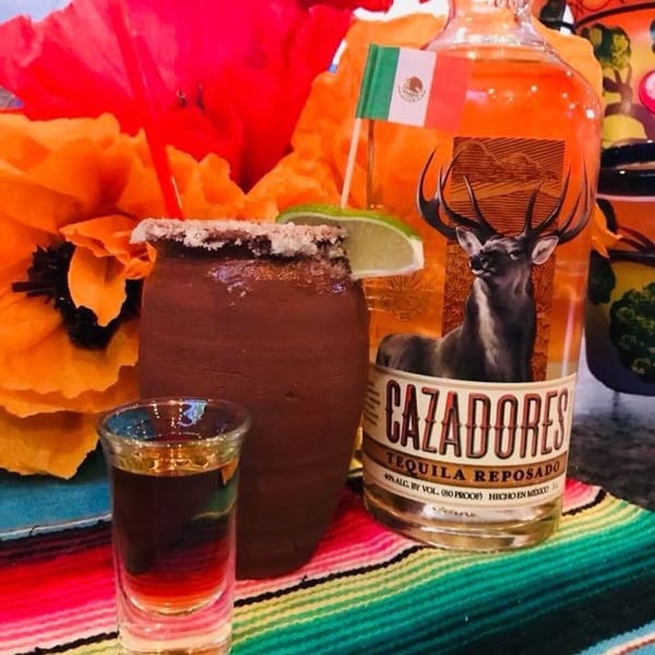 A Cazadores tequila bottle, a clay mug with a lime, and a shot glass on a colorful Mexican serape.