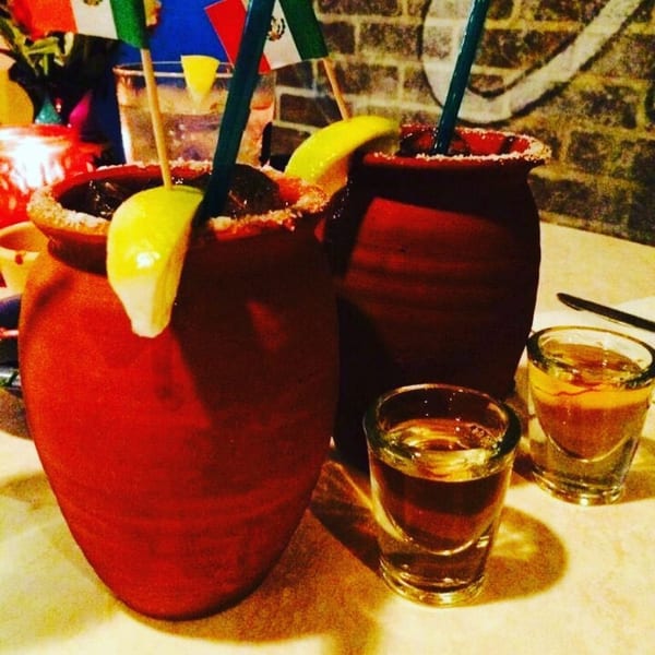 Two red clay cups with drinks, lemon slices, straws, and two shot glasses on a table.