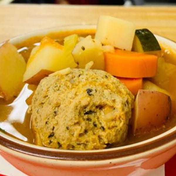 Hearty matzo ball soup with carrots, potatoes, and zucchini in a white bowl on a wooden table.