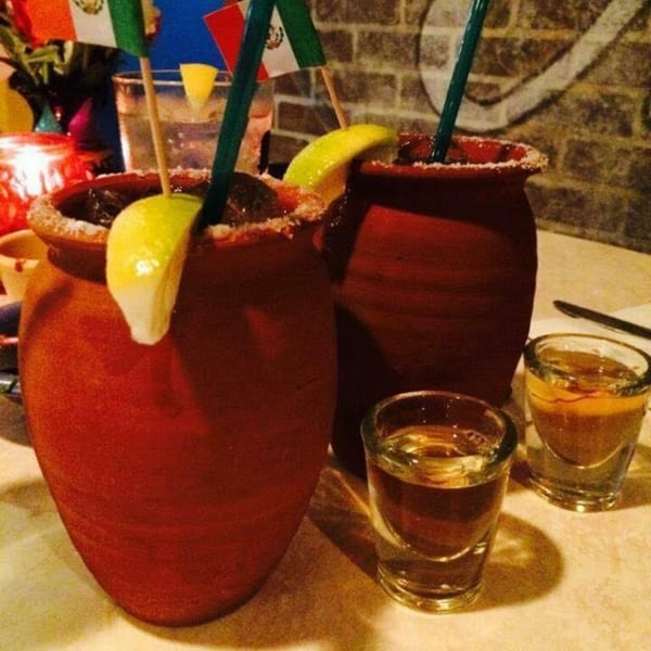 Two large, red clay mugs with margaritas, garnished with lime and salt, alongside two tequila shots.
